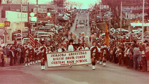 A vintage image of the kilties marching down a street with a banner saying seasons greetings.