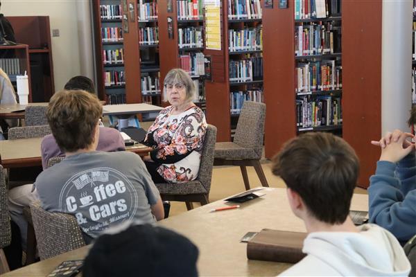 Erika Schwartz sits at a table with several students talking to them in the library.