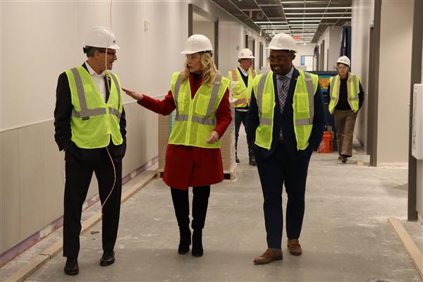 A woman and two men in vests, safety glasses and hard hats walk in an unfinished school construction