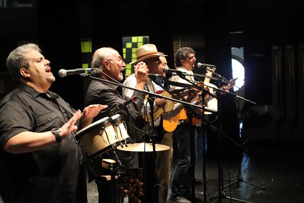 Five man sing into microphone in a line. Each plays an instrument: bongos, maracas and 3 guitars.