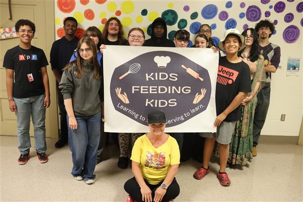 A group of students stands holding a Kids Feeding Kids sign. The teacher sits on the floor in front.