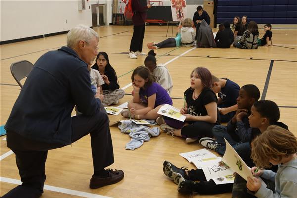 A man kneels to talk to a group of students sitting on a gym floor with paper booklets. 