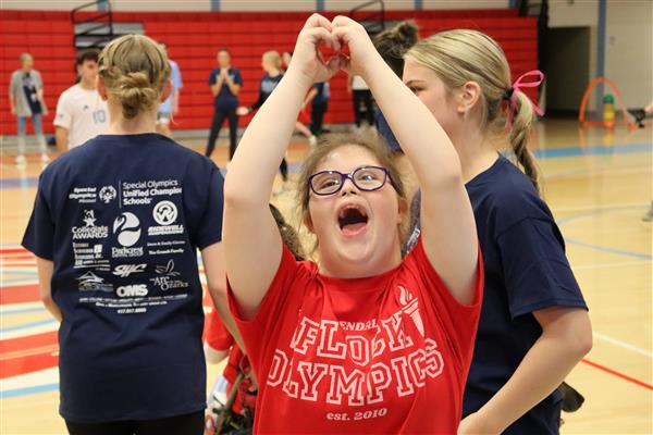  A girl wearing a Flock Olympics shirt in the Glendale gym forms a heart with her hands and smiles.