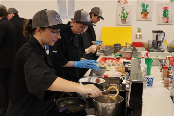 A girl uses tongs to hold an onion ring over hot oil. Behind her students work on burgers.