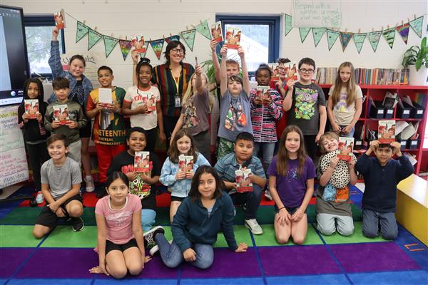 A class of students pose together for the photo. Some student hold up copies of the book Frindle.