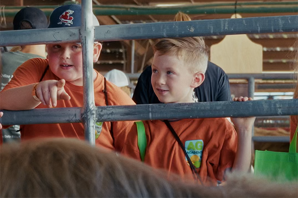 Two boys in AgAcademy t-shirts look at an animal through bars. One boy is pointing at the animal.